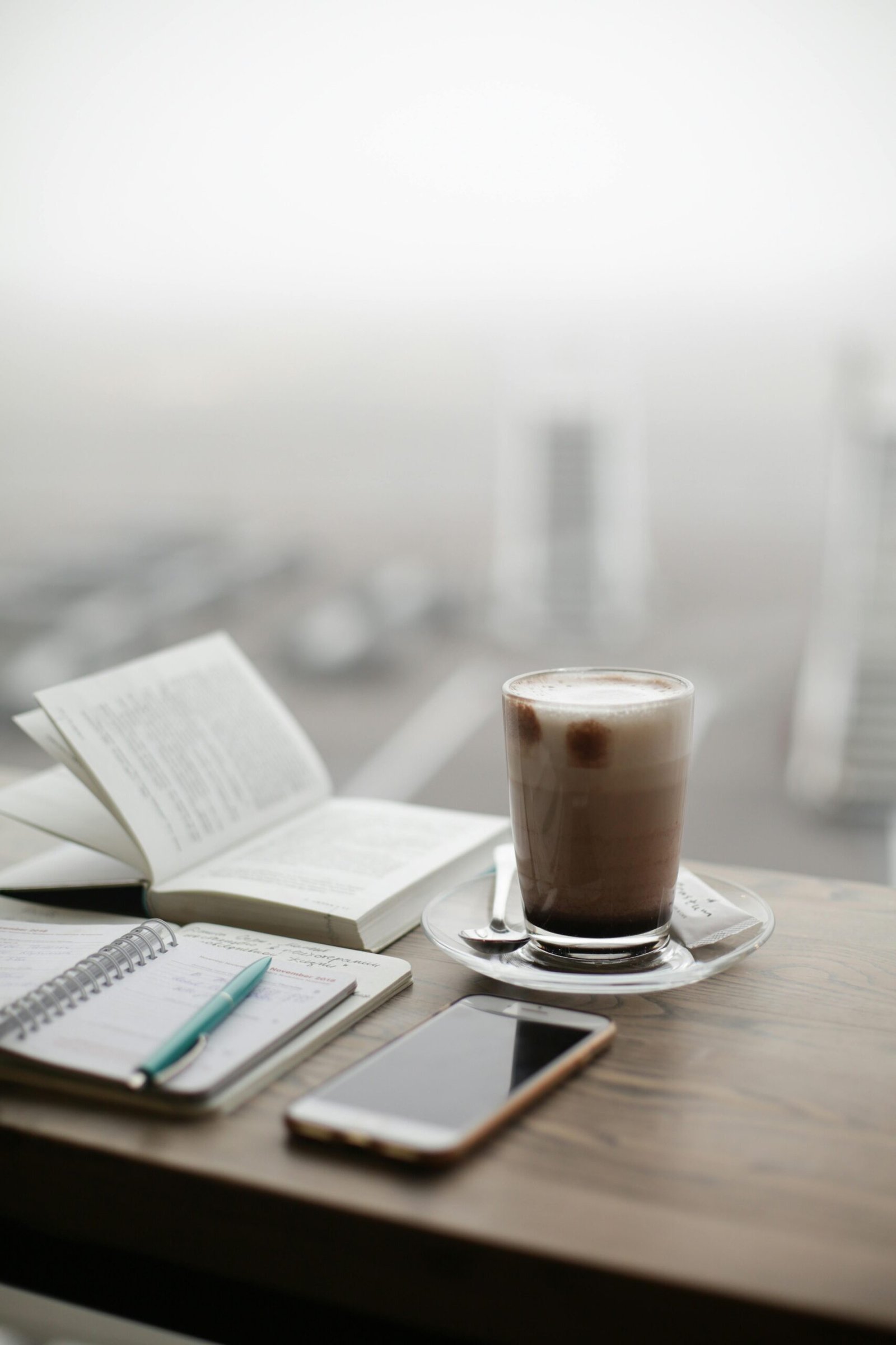 A warm coffee sits beside a notebook and smartphone on a foggy day.